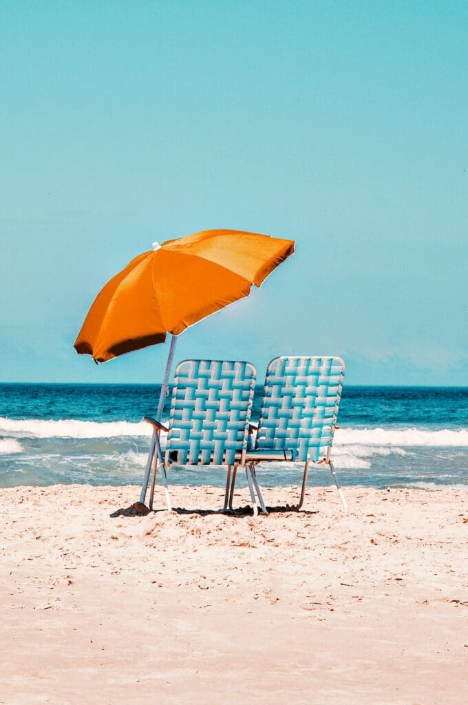 Two chairs on a beach under an orange umbrella.